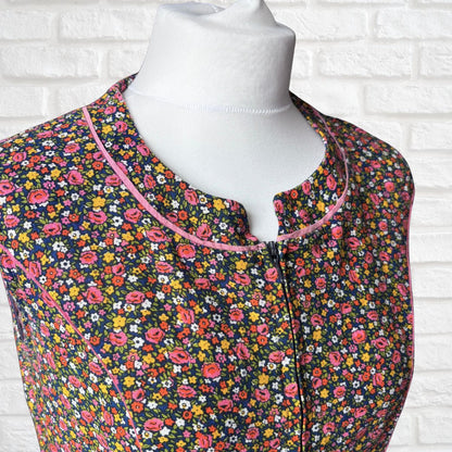 A sleeveless dress with a dense multicolour floral print on a dark background, featuring a pink-trimmed split neckline and a front zipper, displayed on a white mannequin torso against a white brick wall.
