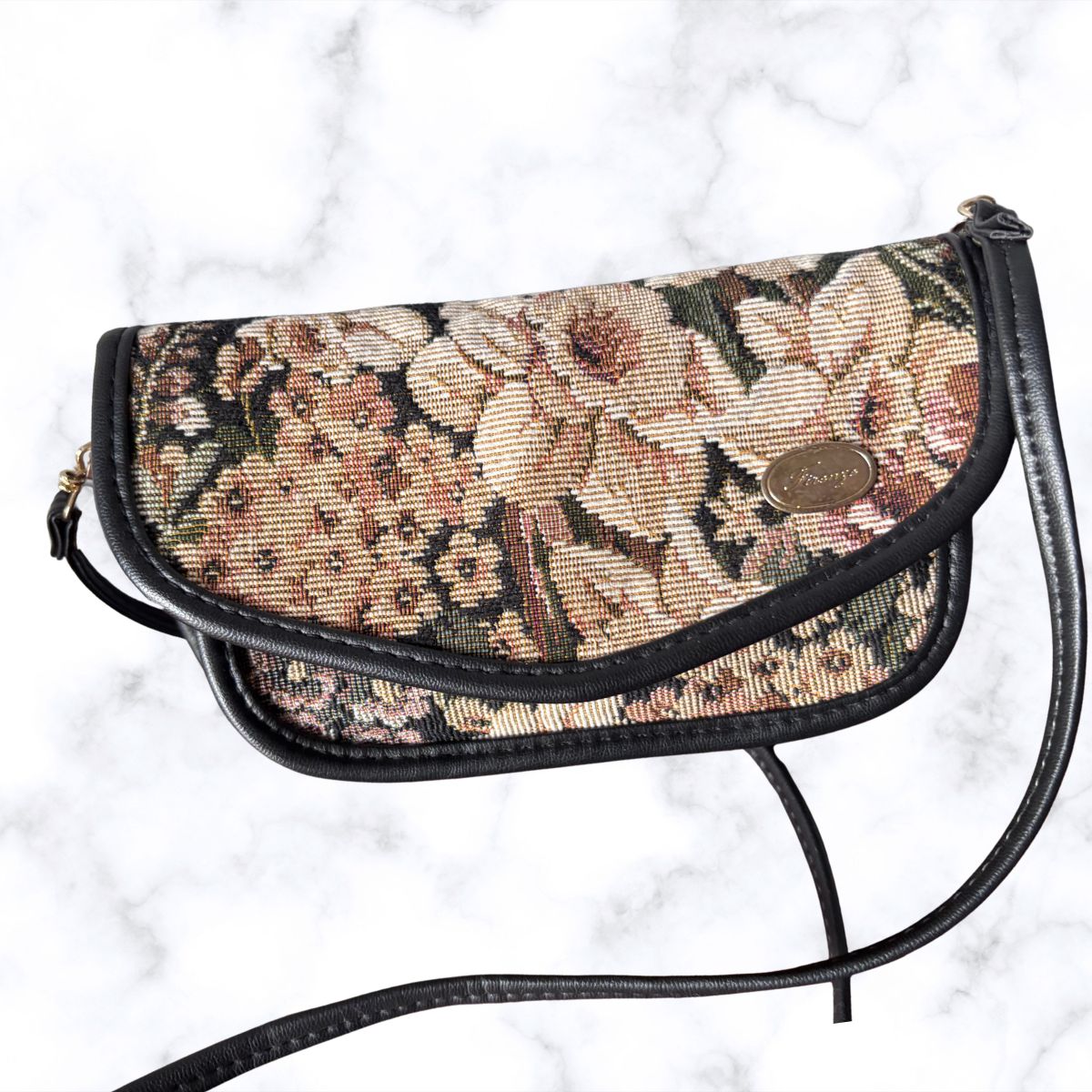 A vintage-style floral tapestry shoulder bag with black leather trim and strap, featuring a gold-tone oval logo medallion on the front flap, set against a white marble background.