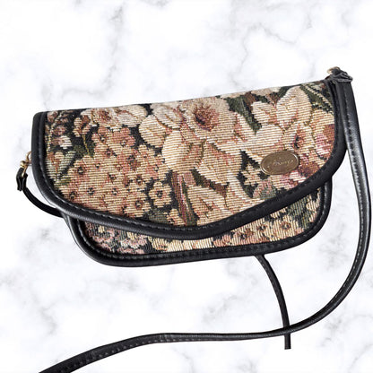 A vintage-style floral tapestry shoulder bag with black leather trim and strap, featuring a gold-tone oval logo medallion on the front flap, set against a white marble background.