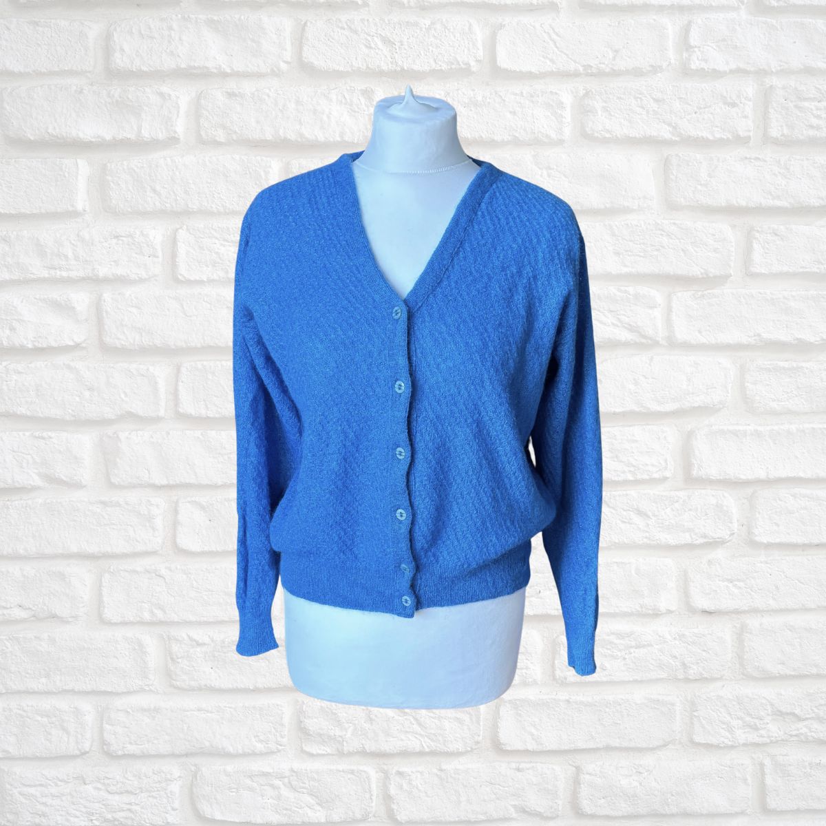 A bright blue textured knit cardigan sweater displayed on a light blue mannequin torso against a white faux-brick background.
