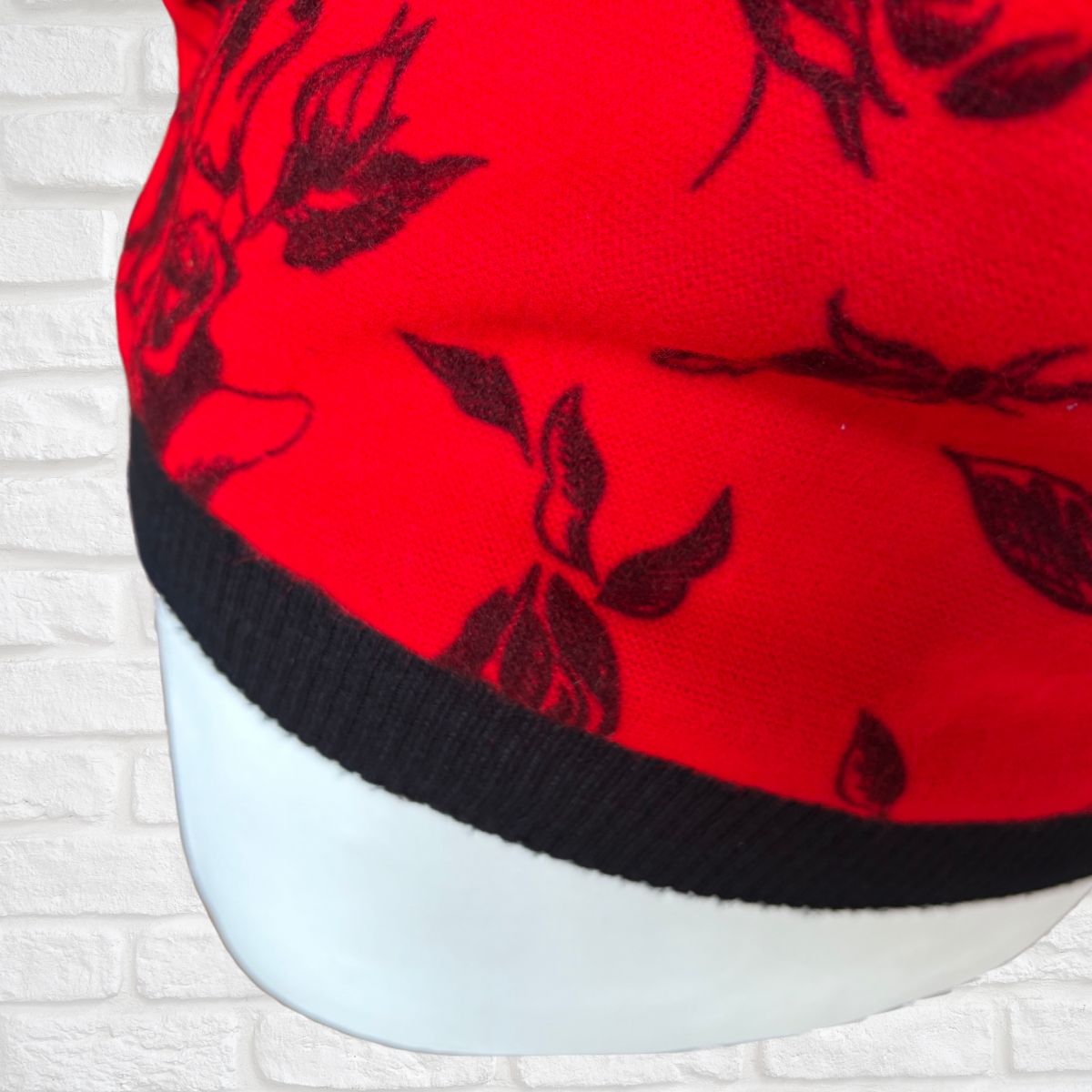 A close-up of a bright red top with a black floral pattern and a black ribbed brim, displayed on a white mannequin head against a white brick wall background.