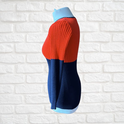 A colorblock ribbed knit sweater displayed on a light blue mannequin torso, set against a white brick wall background. The sweater features a vibrant red upper section, a dark navy blue lower section, and light blue accents at the collar and cuffs.