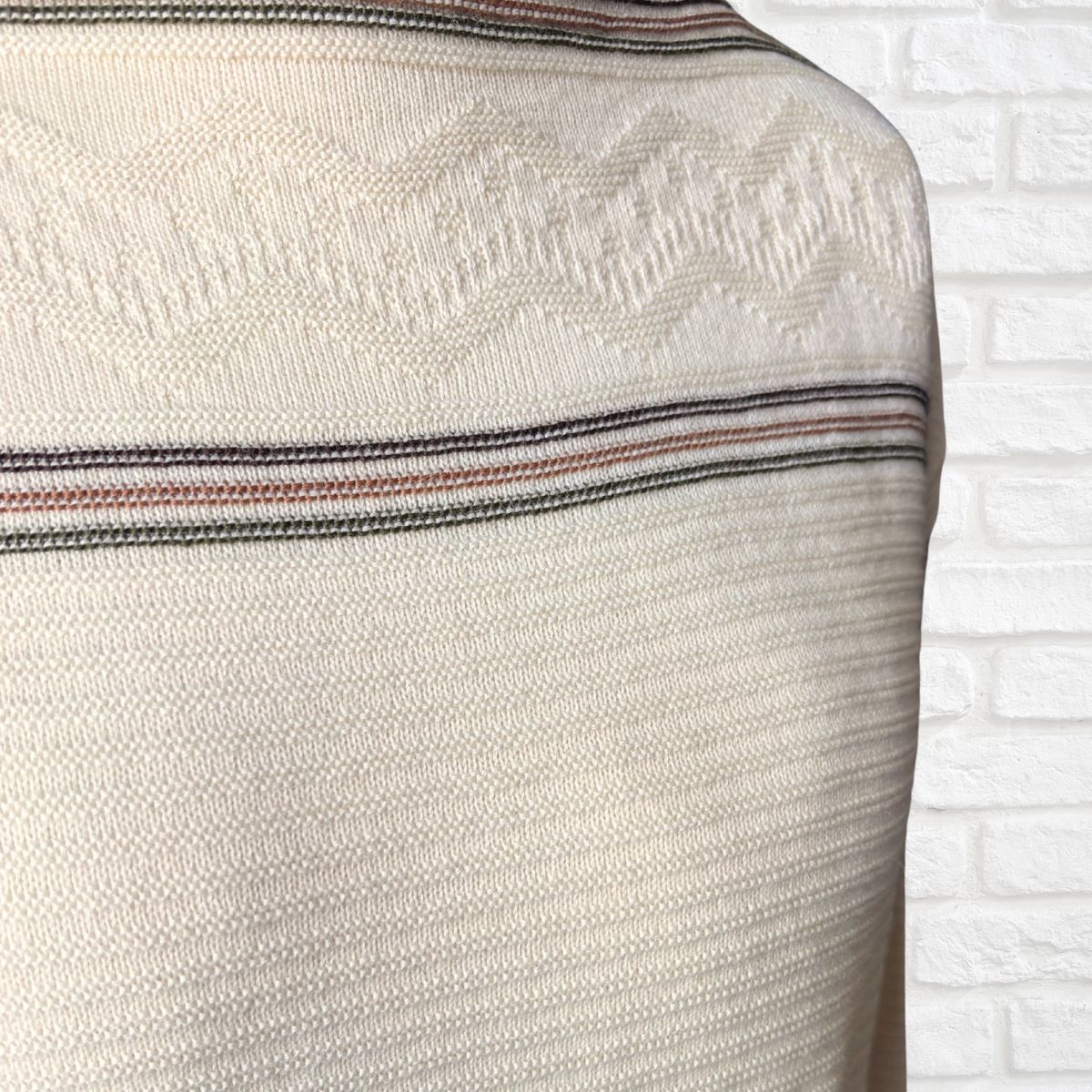 Close-up of a cream-colored knit sweater with a textured geometric pattern and thin horizontal stripes  shown against a white brick wall background.
