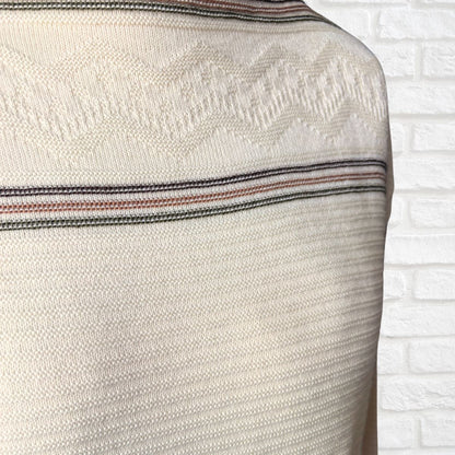 Close-up of a cream-colored knit sweater with a textured geometric pattern and thin horizontal stripes  shown against a white brick wall background.