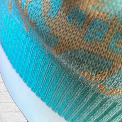 Close-up of a knitted textile with a ribbed turquoise-blue cuff and a tan and blue patterned knit body, shown against a light background.