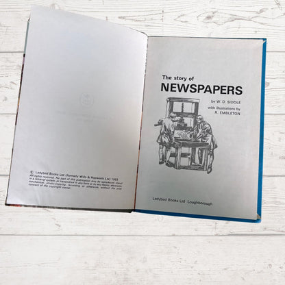 An open book titled 'The story of NEWSPAPERS' laid flat on a white wood surface. The left page shows the interior with printed text and a faint embossed logo, while the right page has a black line drawing of a newspaper press and the title.