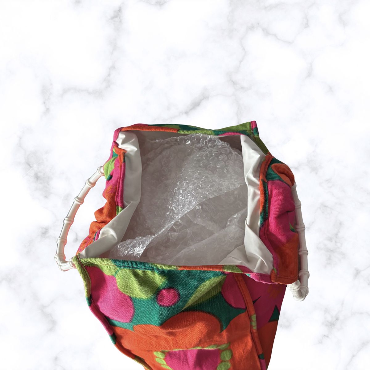 A colorful fabric bag with a vibrant floral pattern in pink, orange, green, and yellow, shown from a top-down view on a white marble background.