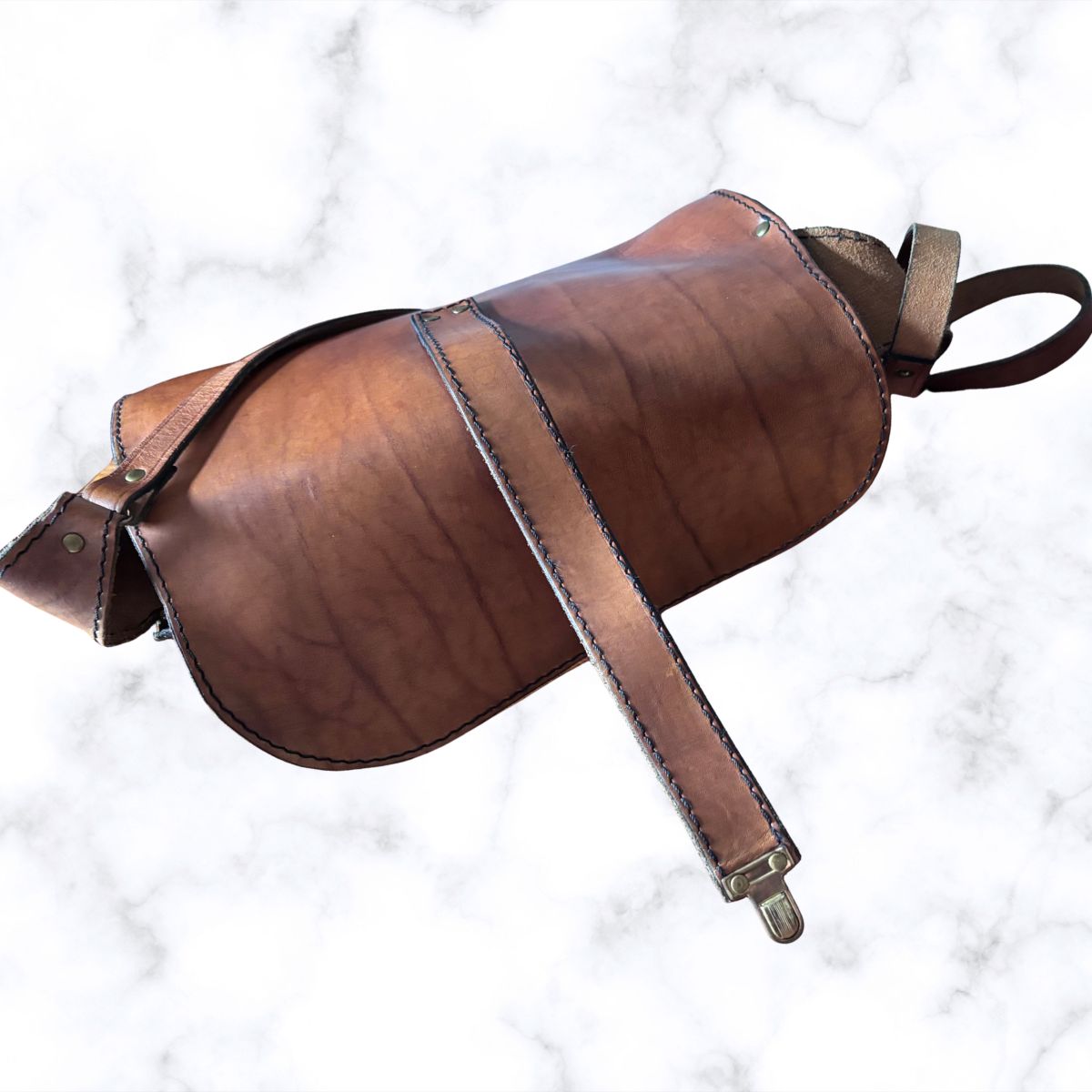 A brown leather saddle bag with a matching leather strap and metal buckle, photographed on a white marble background.