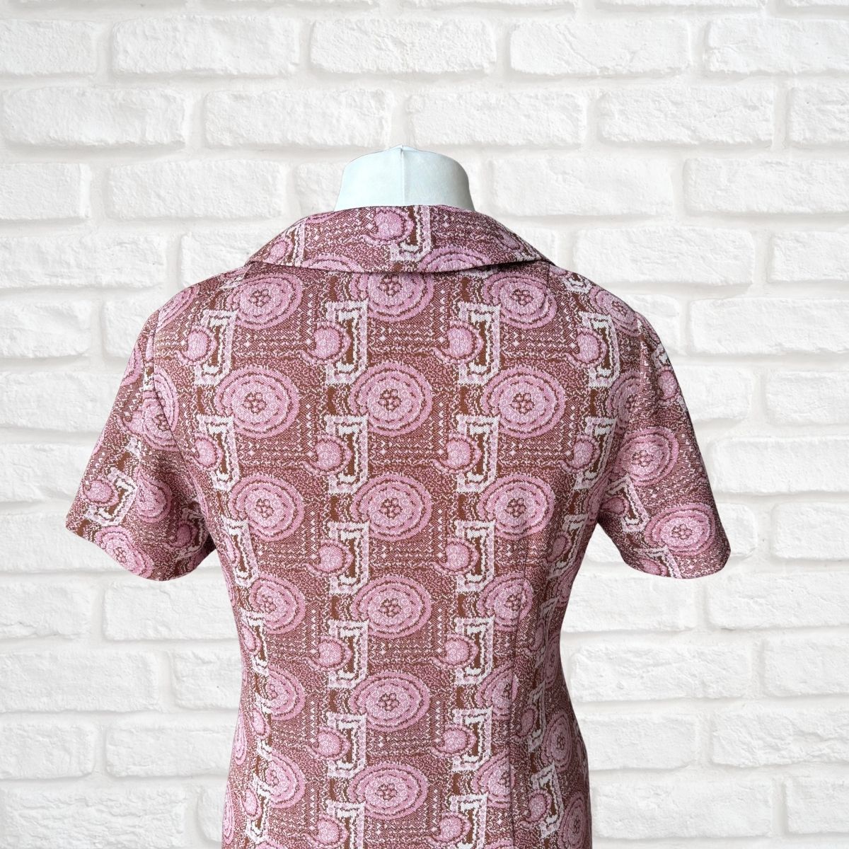 A short-sleeve collared dress displayed on a white mannequin torso, featuring a pink and white geometric pattern with circular medallions, set against a white brick wall background.