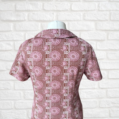 A short-sleeve collared dress displayed on a white mannequin torso, featuring a pink and white geometric pattern with circular medallions, set against a white brick wall background.