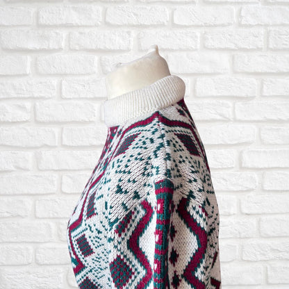 A close-up side view of a knit sweater on a mannequin torso, featuring a white ribbed turtleneck and a geometric Fair Isle pattern in white, teal, and burgundy, set against a white brick wall background.