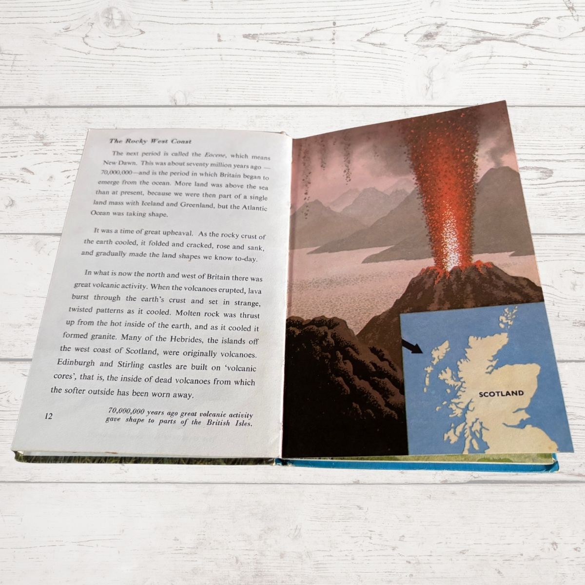 An open children's book spread showing a page about the Rocky West Coast with text on the left and a large illustration of a volcanic mountain on the right, photographed on a white wood surface.
