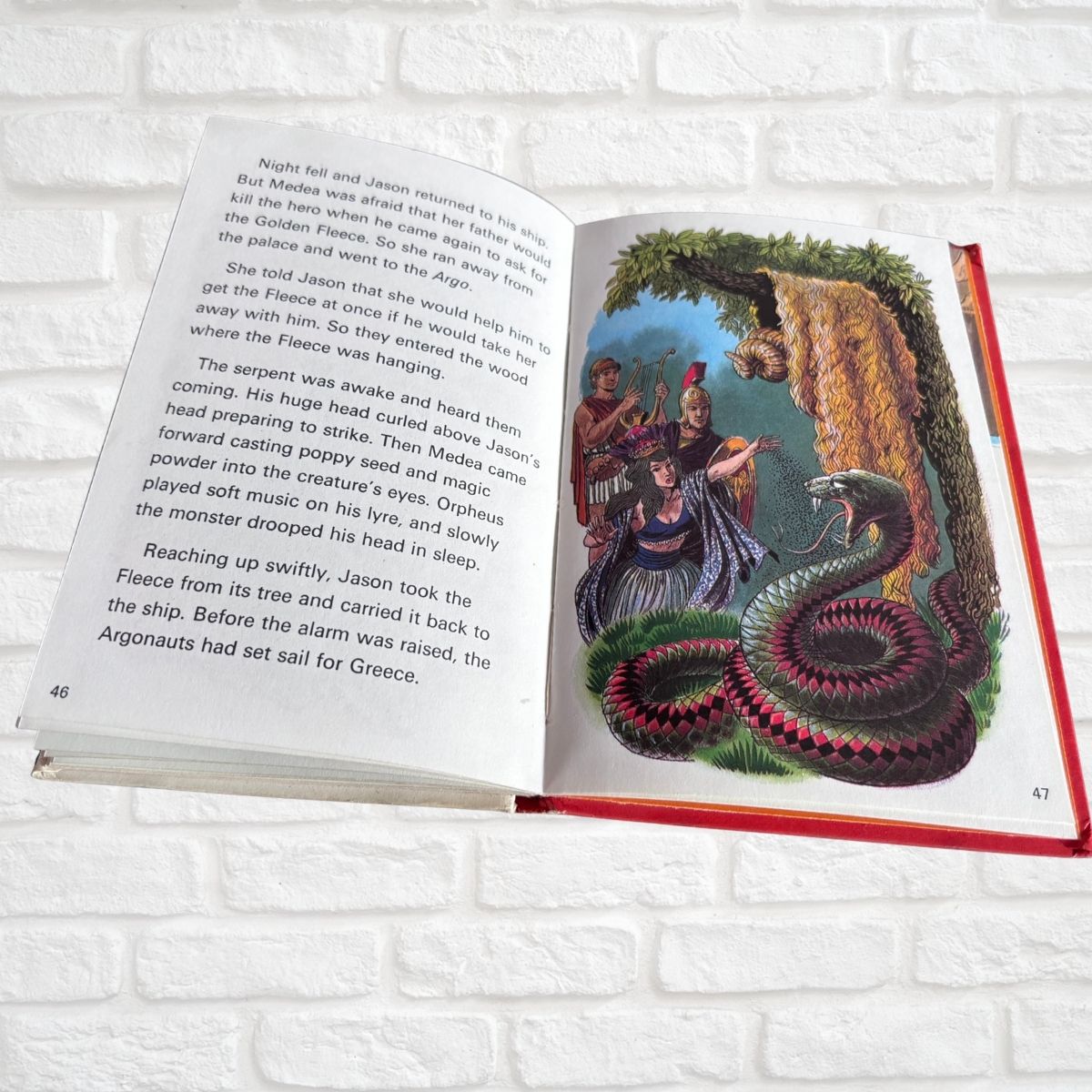 An open children's book lying flat on a white brick background, showing a colorful illustration of a sea serpent around three figures on one page and printed text on the other page.