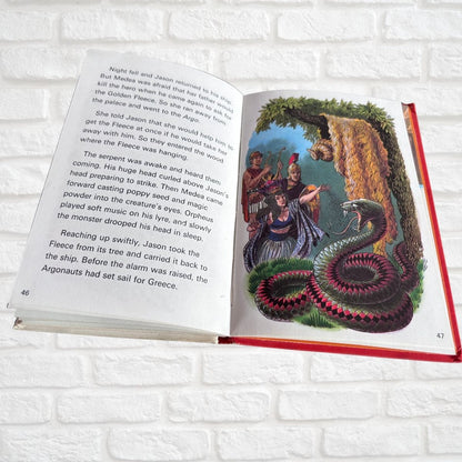 An open children's book lying flat on a white brick background, showing a colorful illustration of a sea serpent around three figures on one page and printed text on the other page.
