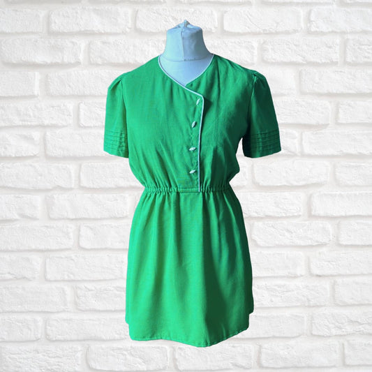 A green short-sleeve mini dress with a V-neckline, buttoned front, and elastic waist, displayed on a light blue mannequin torso against a white brick wall background.