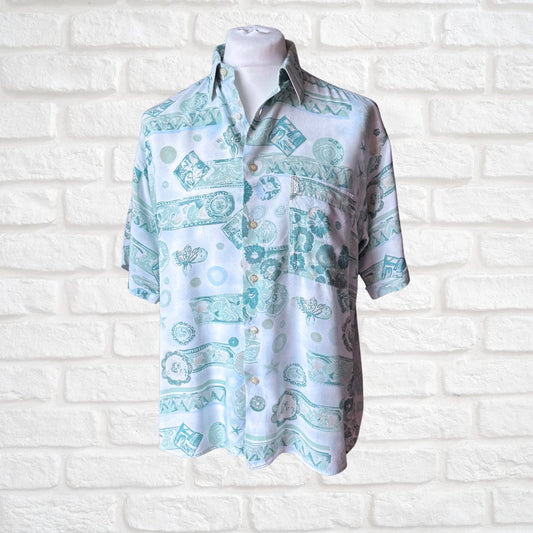 A short-sleeve button-up shirt displayed on a white mannequin torso against a white brick wall. The shirt features a light blue and mint green tropical and geometric pattern on a white base.