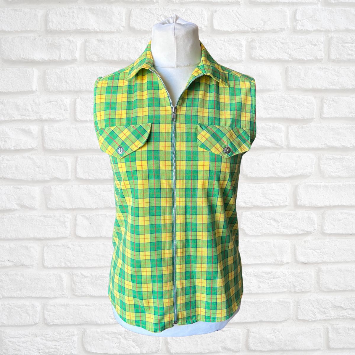 Green and yellow plaid sleeveless shirt on a mannequin against a white brick wall background