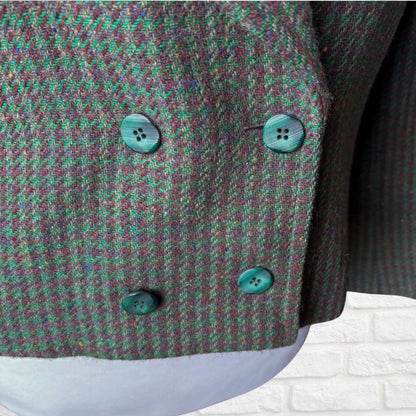 Close-up of a textured tweed jacket or blazer in a green and purple houndstooth pattern, featuring four green buttons, shown on a white brick wall background.