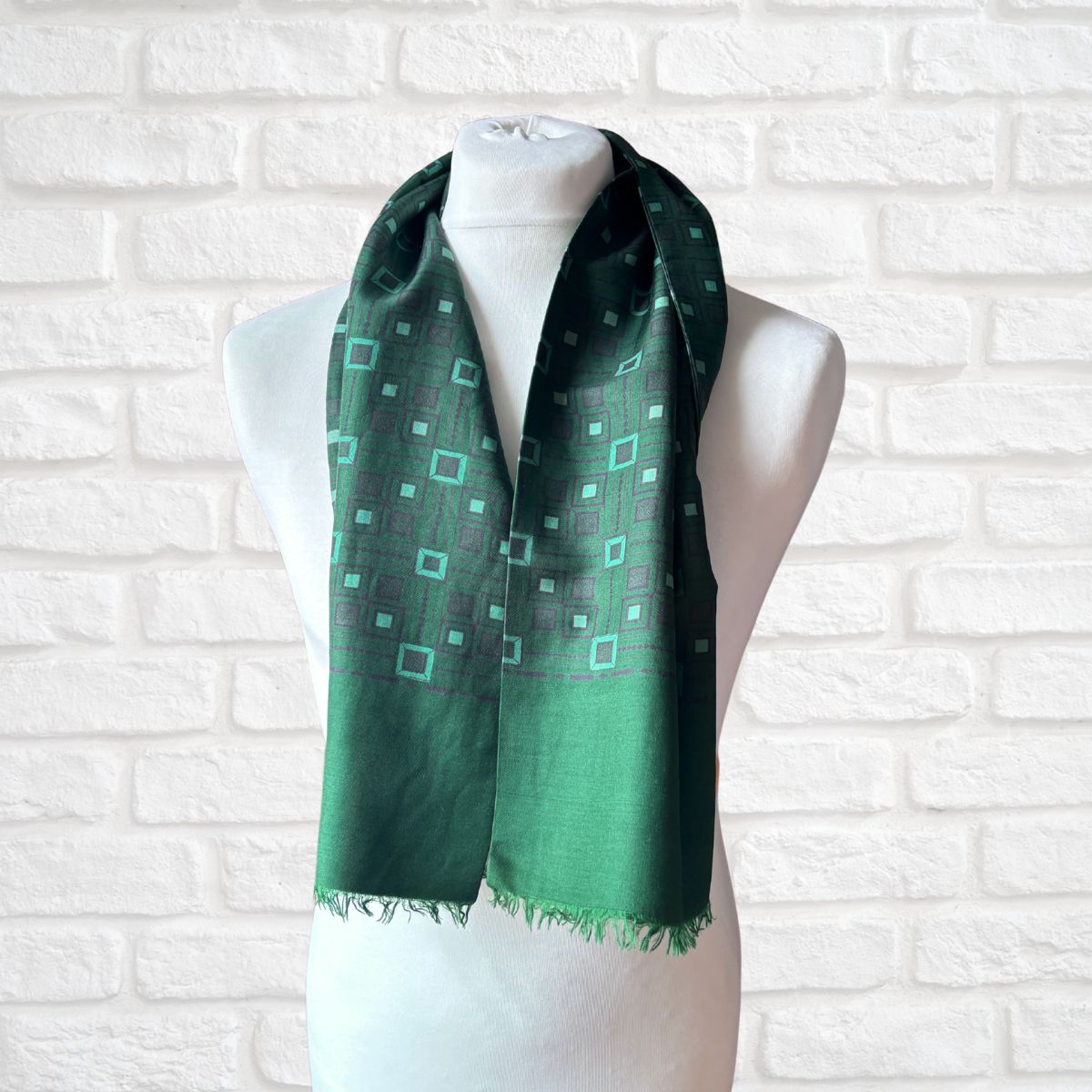 A green scarf with a geometric pattern of light green and purple squares, draped on a white mannequin torso against a white brick wall background.