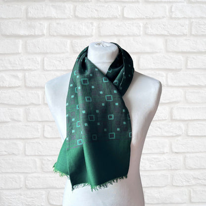 A dark green scarf with a geometric pattern of light green squares, draped on a white mannequin torso against a white brick wall background.