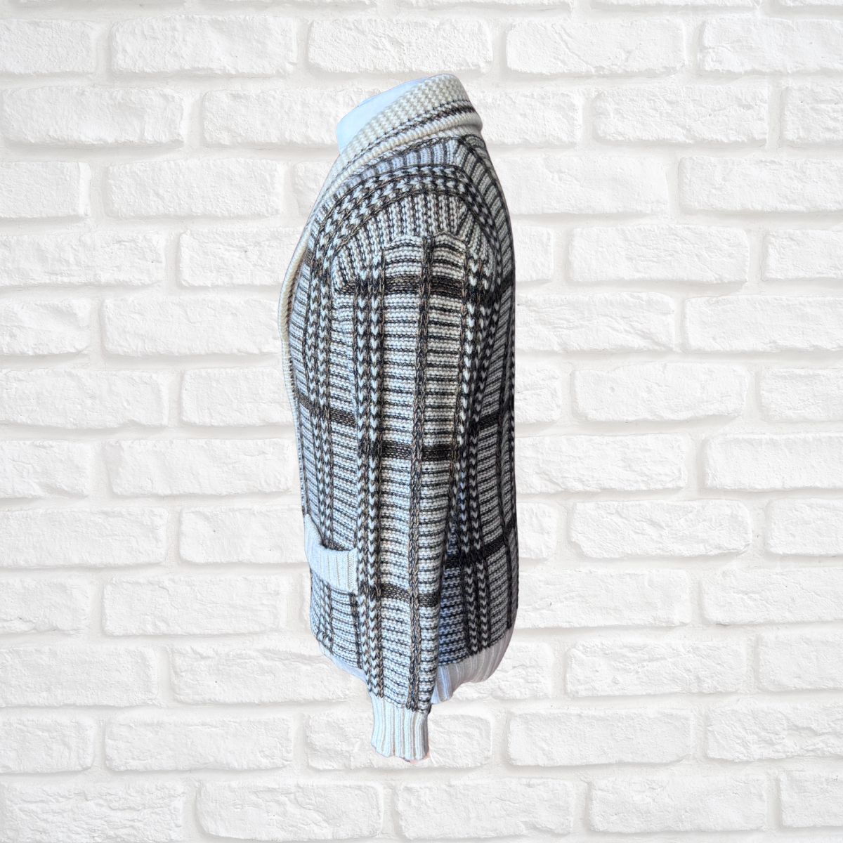 A cream and brown  plaid knit cardigan sweater shown from the side, hanging against a white brick wall background.