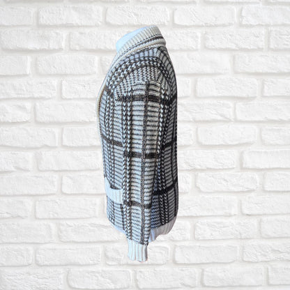 A cream and brown  plaid knit cardigan sweater shown from the side, hanging against a white brick wall background.