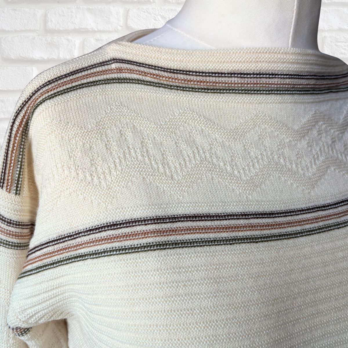 Cream knit sweater with textured chevron patterns and thin horizontal stripes in black, brown, and muted red, displayed on a mannequin torso against a white brick wall background.