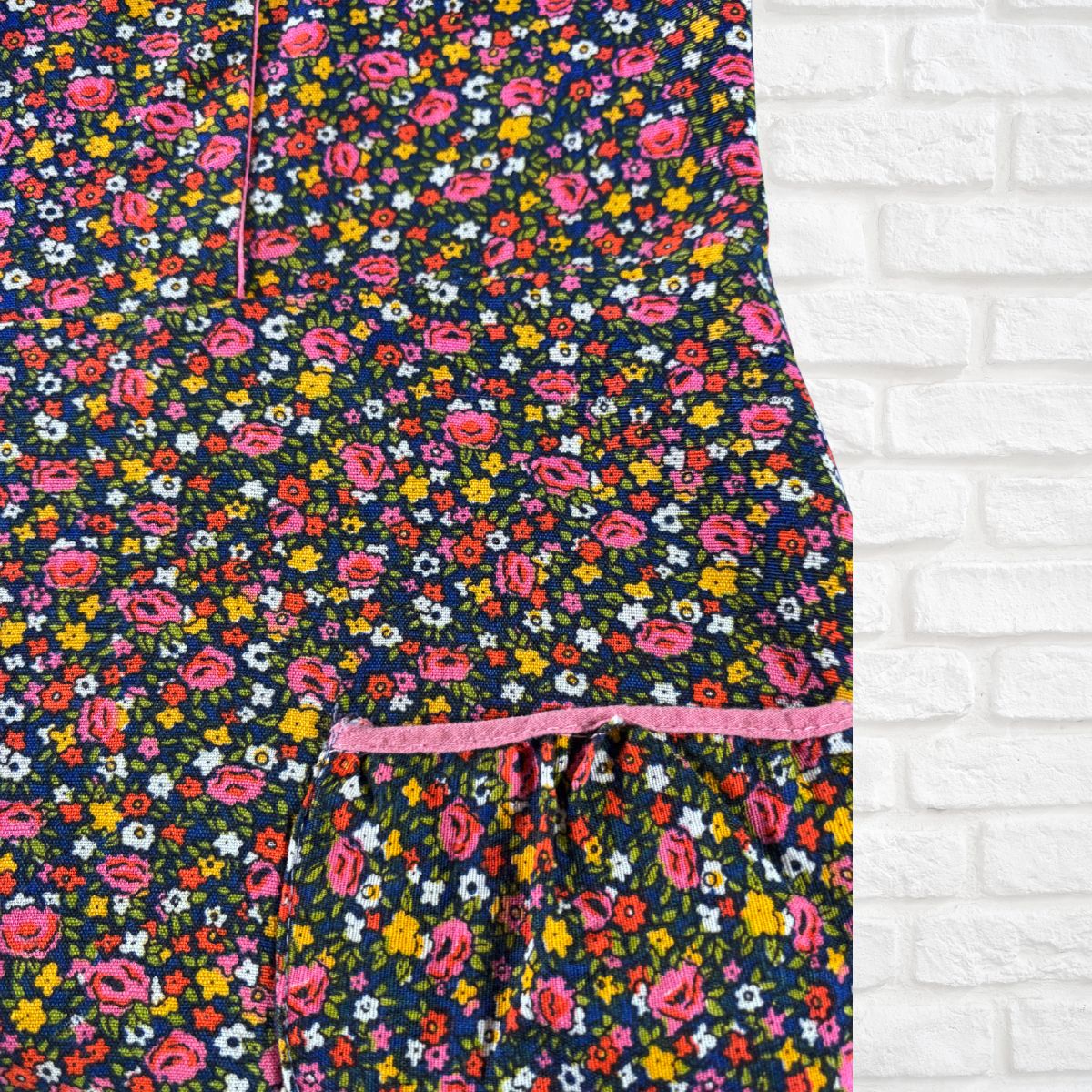 A close-up of a floral-patterned garment, likely a dress or long skirt, featuring a dense print of pink, yellow, and white flowers on a dark navy background, with a pink drawstring waistband, shown against a white brick wall background.