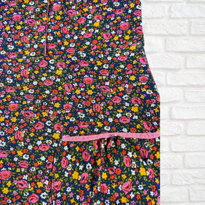 A close-up of a floral-patterned garment, likely a dress or long skirt, featuring a dense print of pink, yellow, and white flowers on a dark navy background, with a pink drawstring waistband, shown against a white brick wall background.