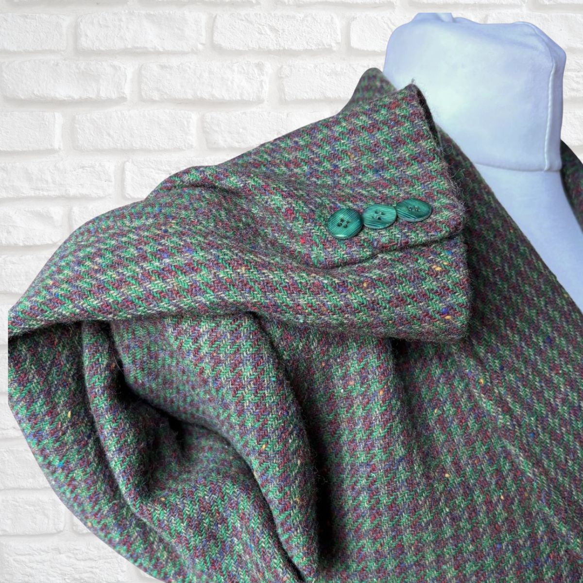 A close-up of a textured tweed blazer  draped on a white mannequin torso, featuring a green and purple brown houndstooth pattern with three green buttons on the collar, set against a white brick wall background.