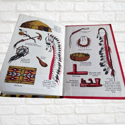 An open vintage educational book displaying illustrations of Native American items such as a war bonnet, flintlock pistol, and a pipe, set against a white brick wall background.