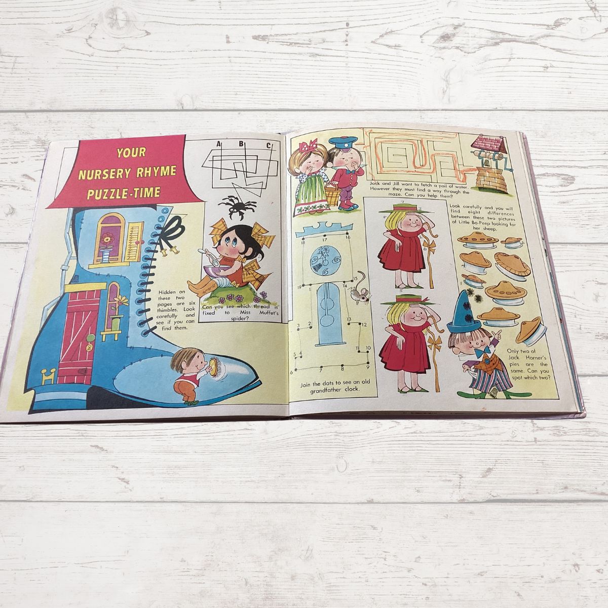 An open vintage children's puzzle book titled 'Your Nursery Rhyme Puzzle Time' laid flat on a white painted wood surface. The pages feature colorful cartoon illustrations, including a blue house, a spider, a door, and whimsical characters, with some printed puzzles and rhymes.