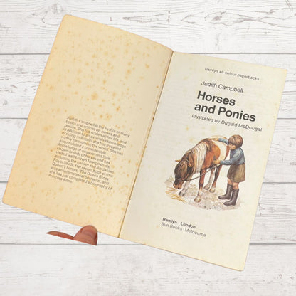 An open vintage children's book titled 'Horses and Ponies' by Judith Campbell, showing aged cream pages with printed text and an illustration of a child feeding a horse, photographed on a white wood background.
