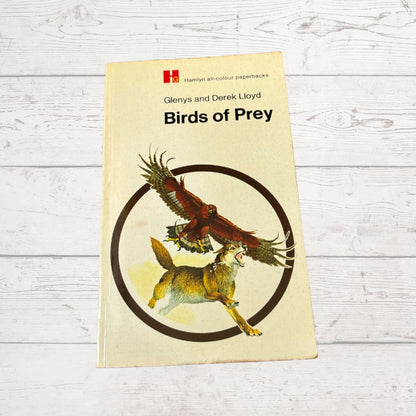 A paperback book titled 'Birds of Prey' by Glenys and Derek Lloyd, published by Hamlyn, lying flat on a white wood-plank surface. The cover is cream-colored with an illustration of a brown bird and a fox inside a dark circle.