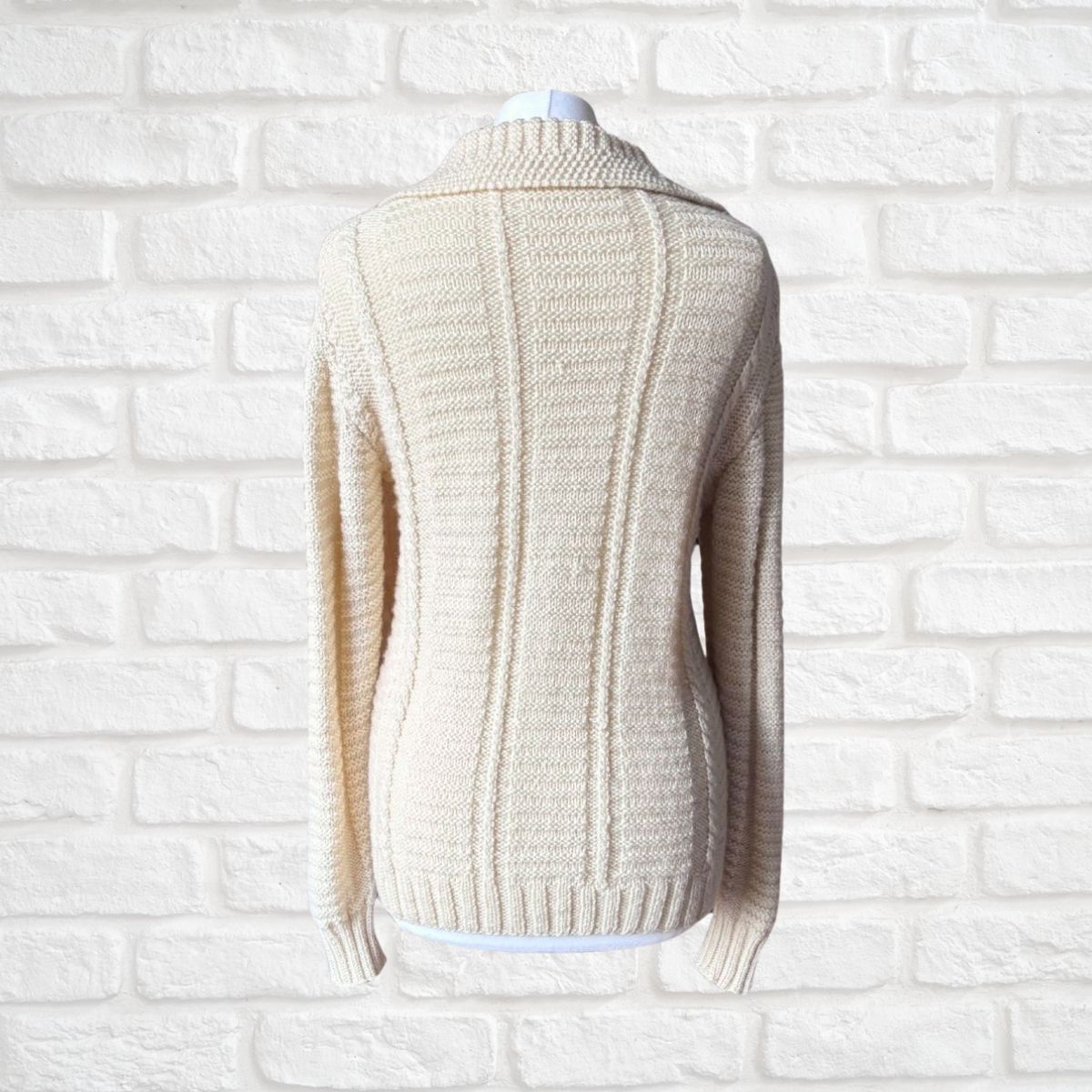 A cream-colored cable-knit sweater displayed on a white mannequin torso against a white brick wall background.