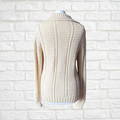 A cream-colored cable-knit sweater displayed on a white mannequin torso against a white brick wall background.