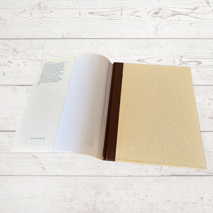 An open book o lying on a white wood plank surface, with cream-colored pages, a dark brown spine, and a large blank beige cover on the right side.