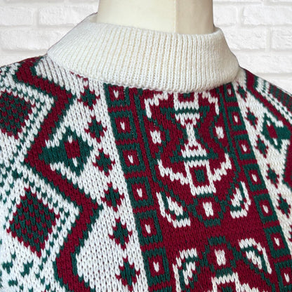 A close-up of a knit sweater on a mannequin torso, featuring a festive geometric pattern in deep red, dark green, and white. The sweater has a ribbed cream-colored mock neck collar.