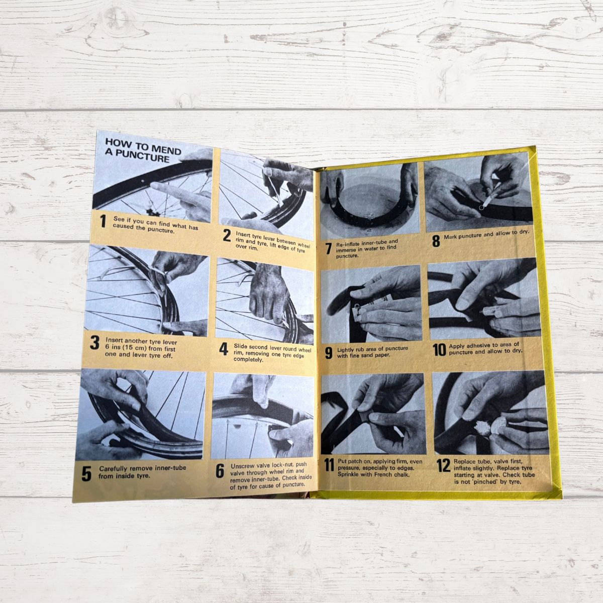 An open instruction book with 12 numbered steps on how to mend a puncture, laid flat on a light whitewashed wooden surface.