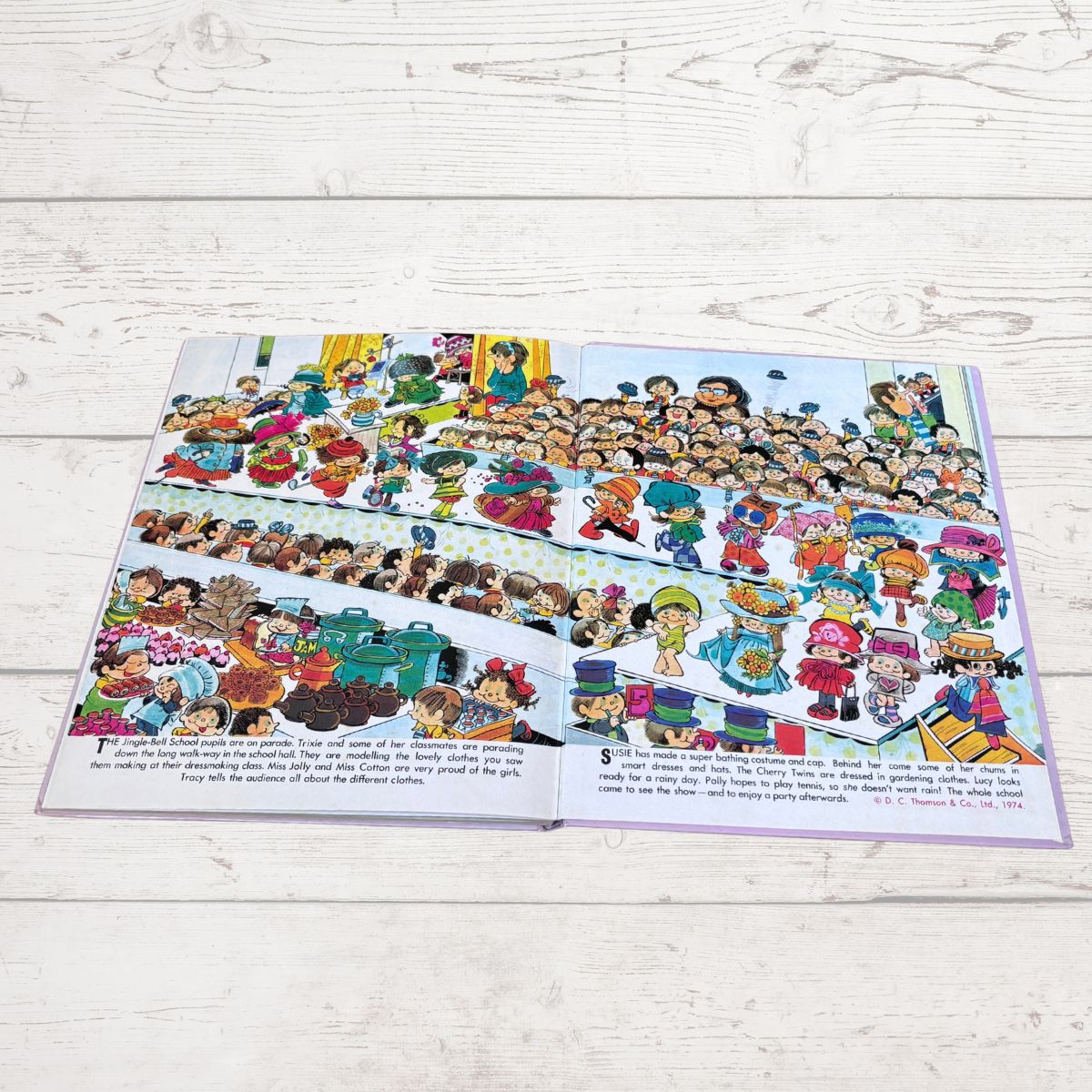 An open children's book laid flat on a light whitewashed wood surface, showing a colorful illustrated scene with many cartoon children and small text blocks along the bottom.
