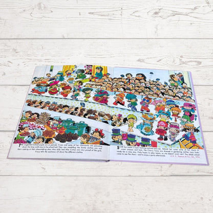 An open children's book laid flat on a light whitewashed wood surface, showing a colorful illustrated scene with many cartoon children and small text blocks along the bottom.