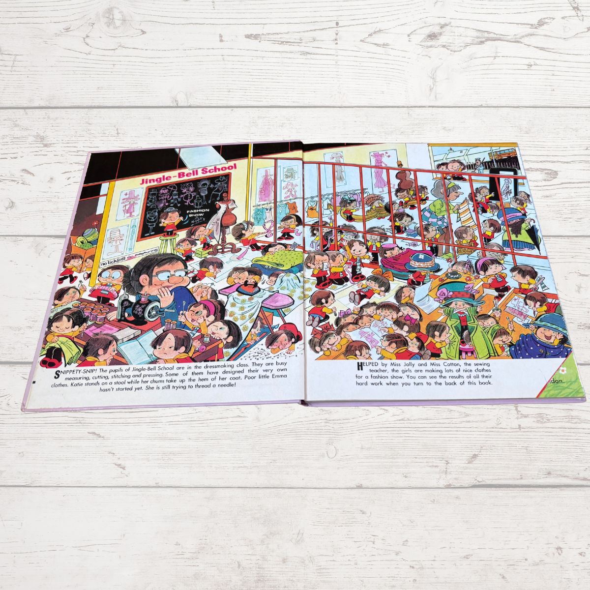 An open children's picture book spread on a white wood plank surface, showing a colorful cartoon illustration of a school scene with many children and adults.