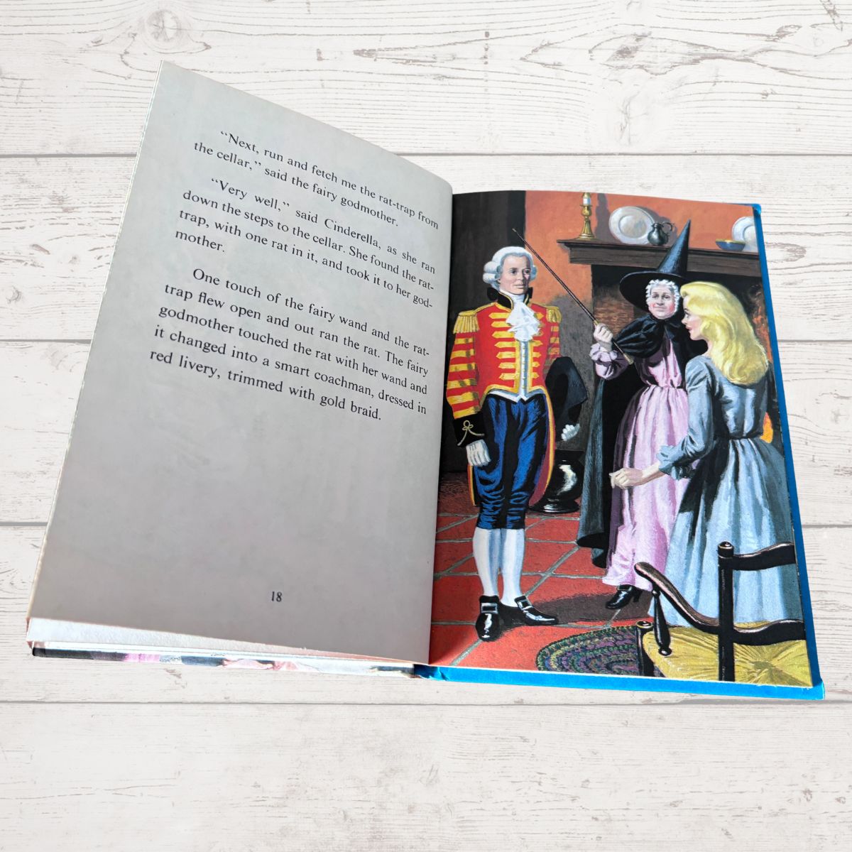 An open children's book lying on a light wood surface, showing a printed story page with text on the left and an illustrated scene on the right featuring a fairy godmother, a child, and a rat.