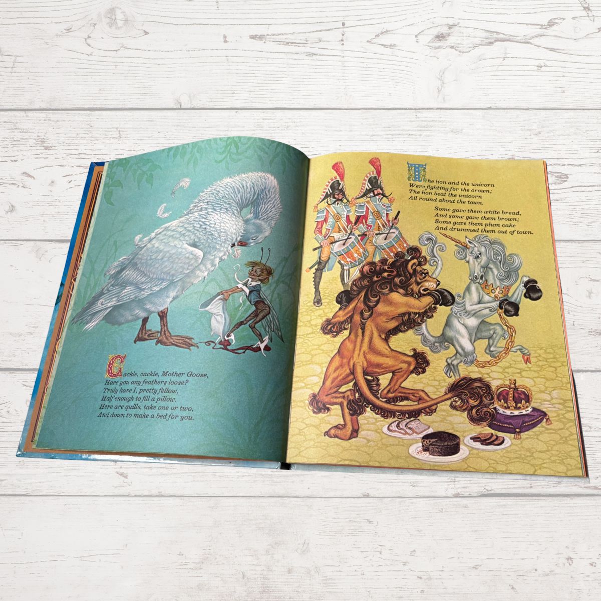 An open children's book spread on a white wood surface, showing illustrated pages with a goose, a lion, a unicorn, and a crown. No visible brand text is legible.