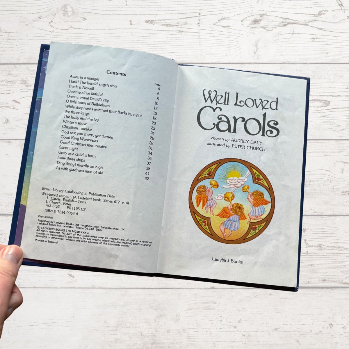Open book titled 'Well Loved Carols' with a cover illustration of children singing, held against a light wooden background.