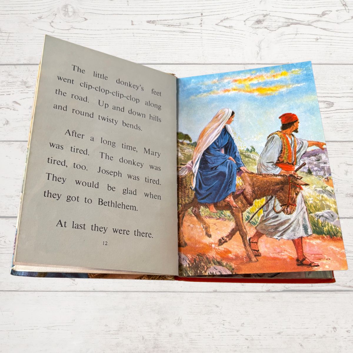 Open book with a colorful illustration of Mary and Joseph on a donkey, set against a white wooden background.