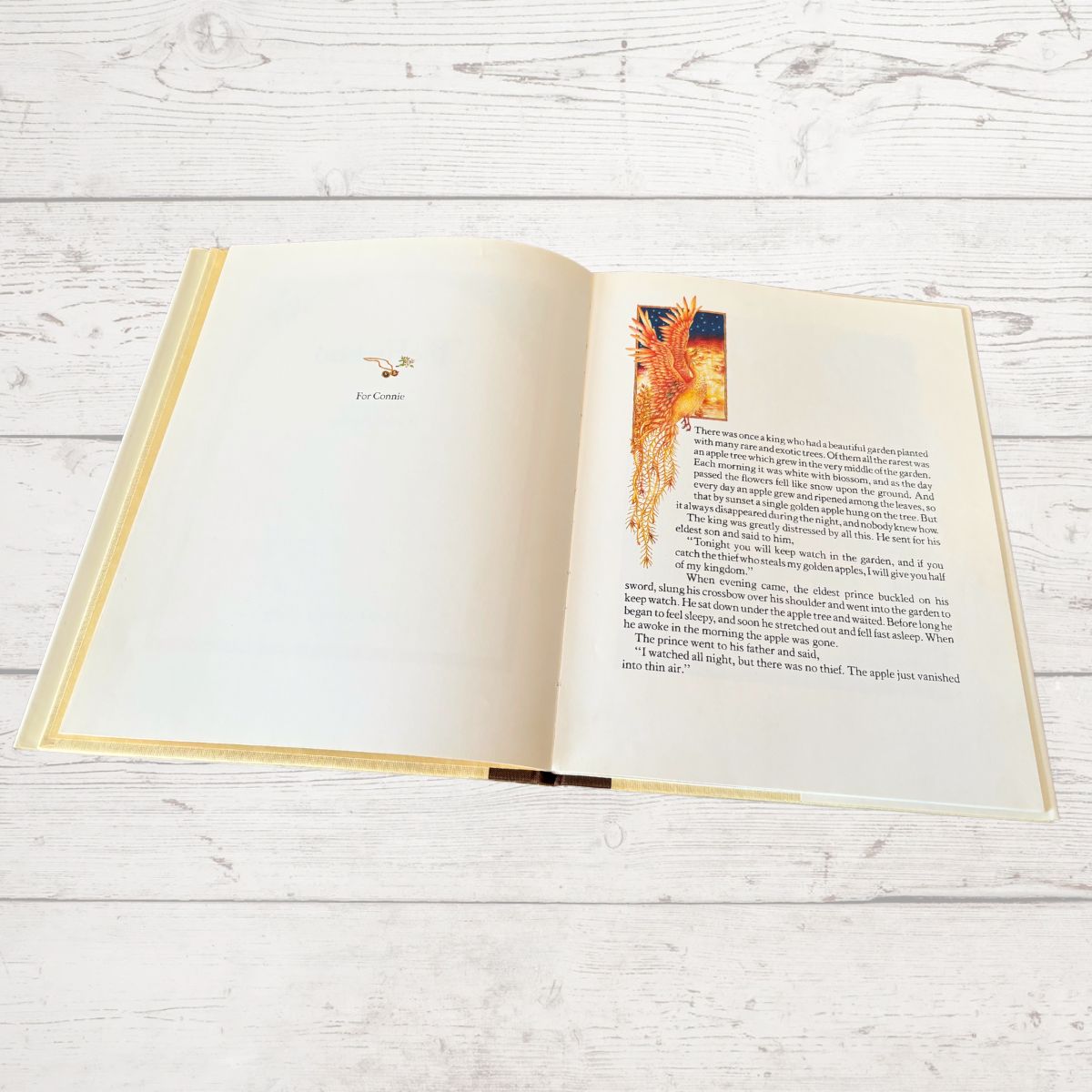 An open hardcover book with cream-colored pages, featuring a small illustration and a larger decorative text block on the right page, photographed on a light whitewashed wood surface.