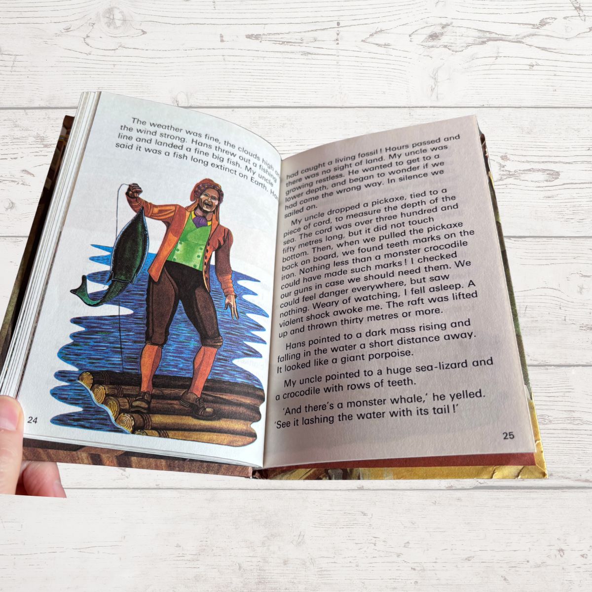 An open children's book being held by a hand, showing two printed pages with text and a colorful illustration of a child sailor on a ship. The book is photographed on a light whitewashed wood surface.