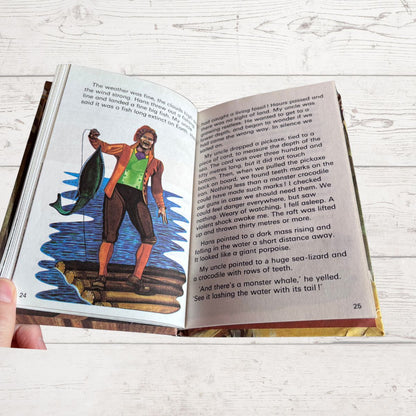 An open children's book being held by a hand, showing two printed pages with text and a colorful illustration of a child sailor on a ship. The book is photographed on a light whitewashed wood surface.