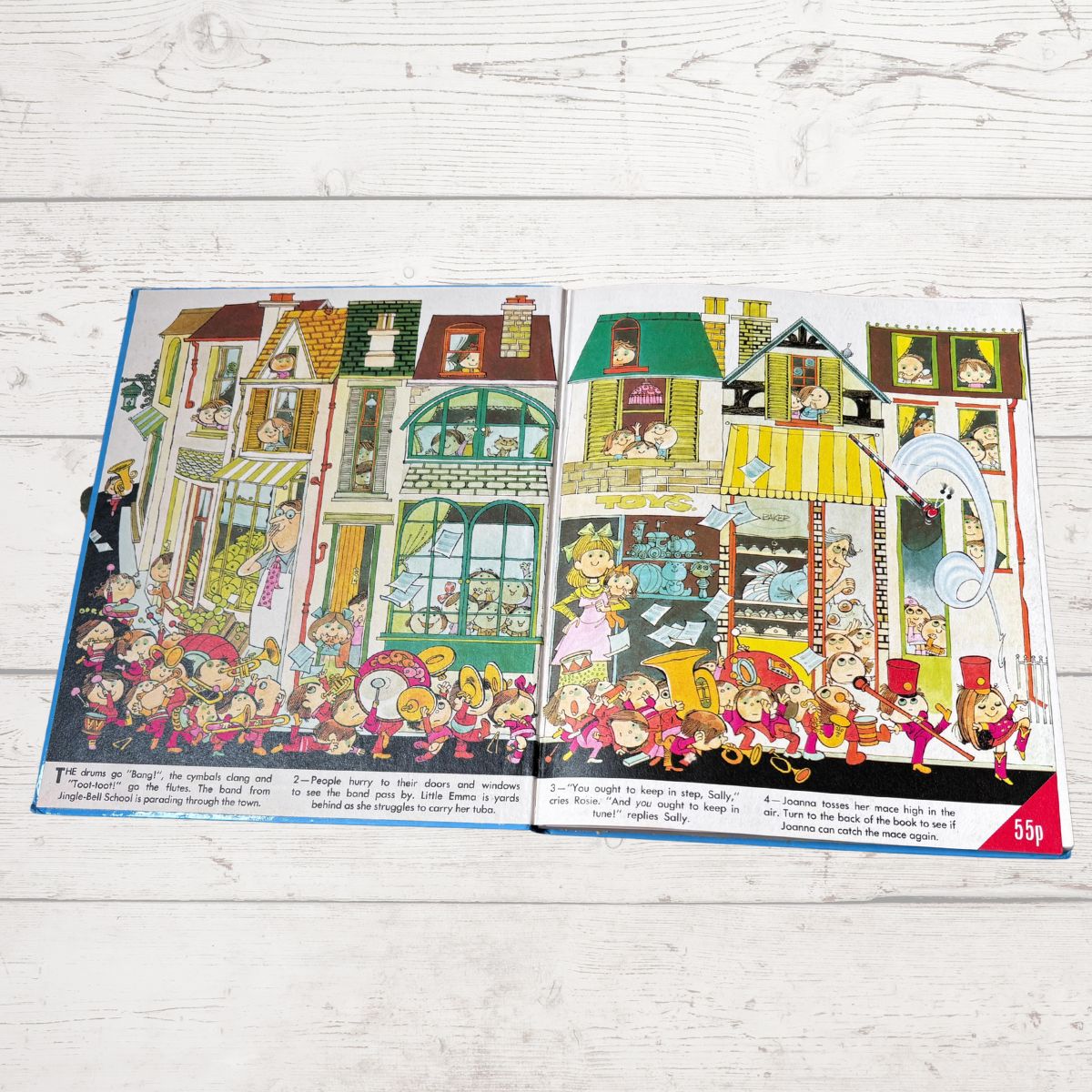 An open illustrated book spread laid flat on a white wood surface, showing a colorful cartoon street scene with many characters and storefronts. The book pages are the main product, and no person or real environment is present beyond the tabletop background.